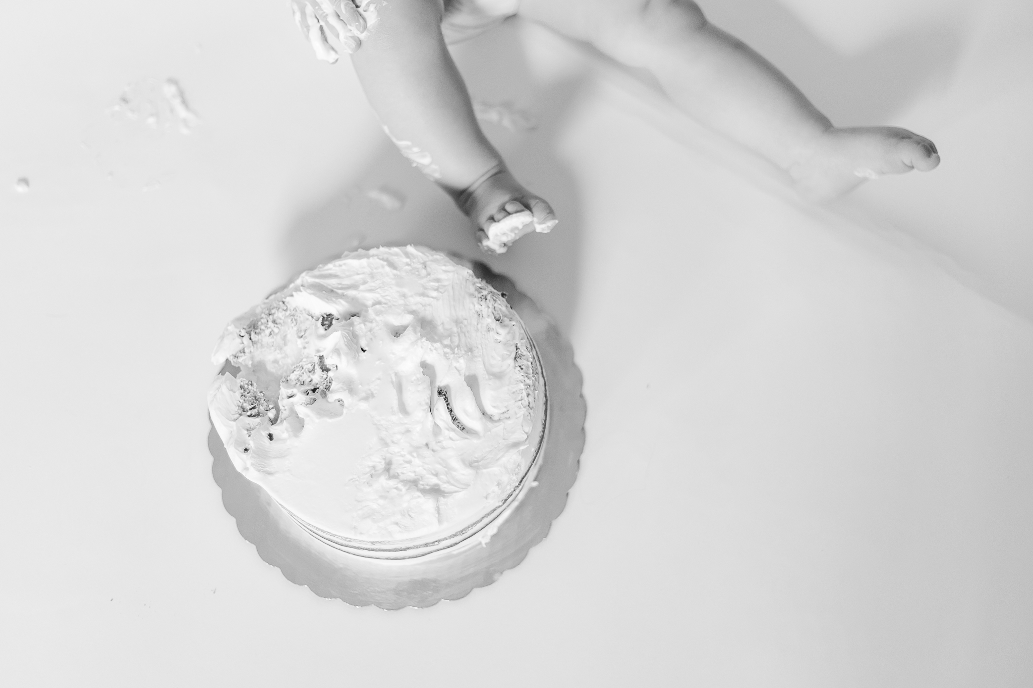 Black and white overhead detail from a one year milestone cake smash session, capturing baby feet and hands reaching into a simple birthday cake on a white backdrop.