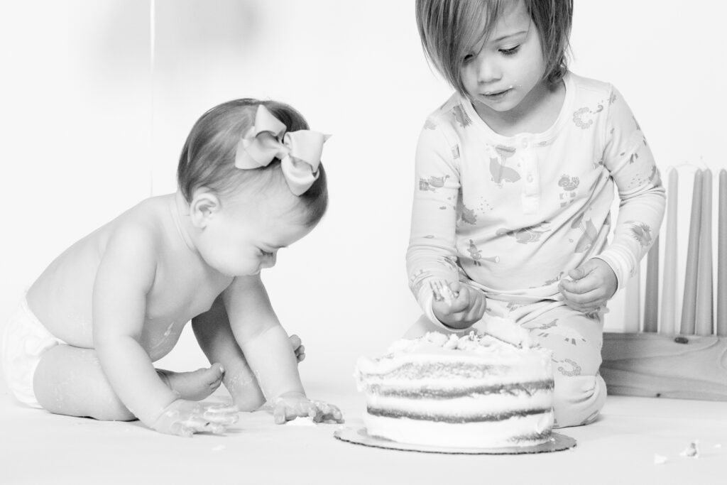 Black and white sibling moment during a one year milestone cake smash, capturing big brother Ivan laughing with baby sister Wren beneath a simple first birthday balloon on a white backdrop.