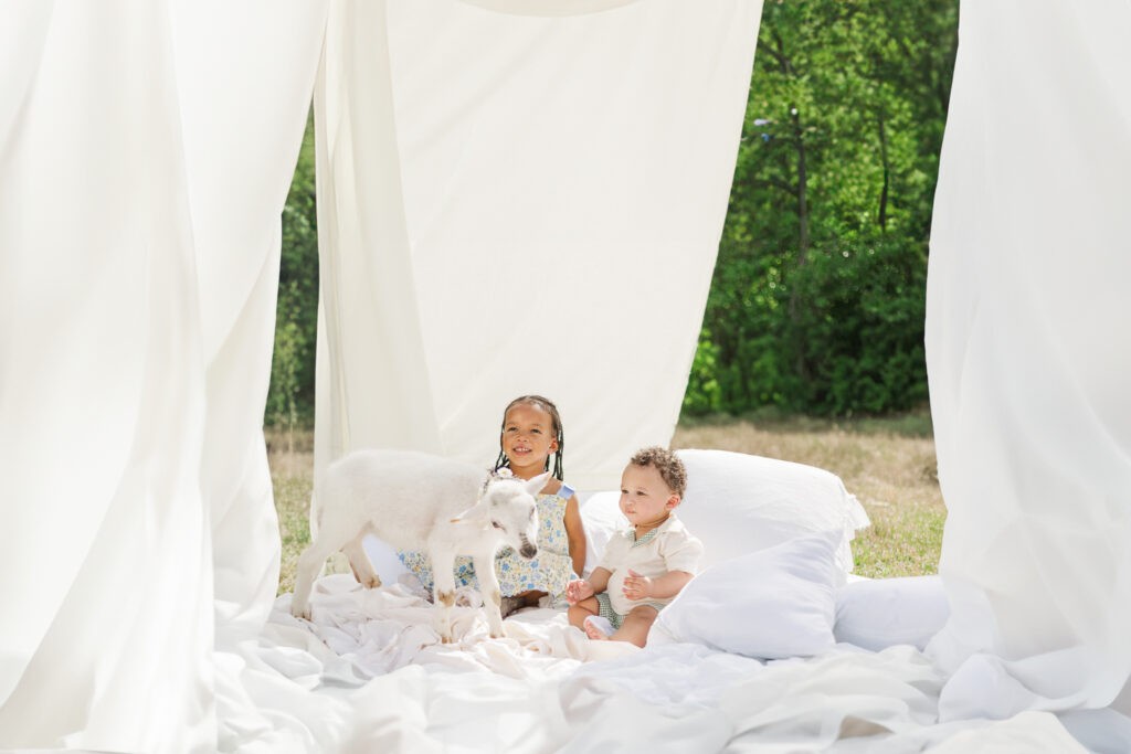 Two young children sitting on soft white linens outdoors with a gentle lamb during an Easter mini session, surrounded by flowing white fabric and spring greenery in Alabama.